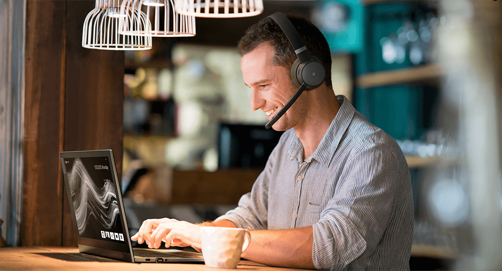 Uomo seduto e sorridente in un bar mentre utilizza un Notebook e con indosso delle cuffie wireless con microfono.