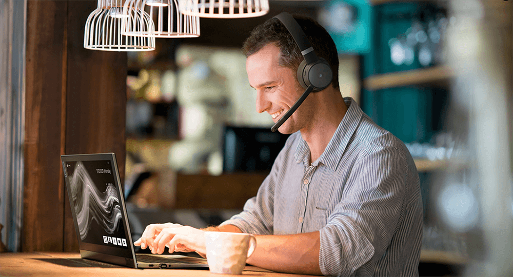 Uomo seduto e sorridente in un bar mentre utilizza un Notebook e con indosso delle cuffie wireless con microfono. 